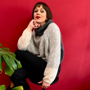 Op de afbeelding: Een vrouw draagt een grijze en witte gebreide trui met een zwarte turtleneck. Ze zit op een kruk met een plant op de voorgrond.