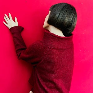 May include: A woman with short, dark hair is wearing a maroon sweater and orange trousers. She is standing in front of a bright red wall and has her hand on the wall.