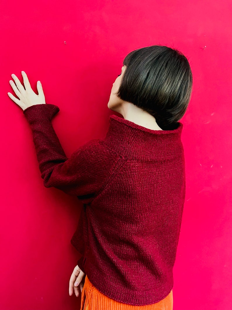 May include: A woman with short, dark hair is wearing a maroon sweater and orange trousers. She is standing in front of a bright red wall and has her hand on the wall.
