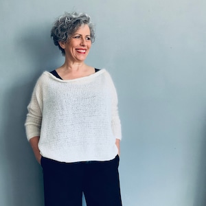 Könnte beinhalten: Eine Frau mit kurzen, grauen, lockigen Haaren trägt einen weißen, gestrickten Pullover mit lockerer Passform und einem weiten Ausschnitt. Sie trägt eine schwarze Hose und hat einen neutralen Gesichtsausdruck.