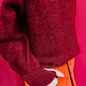 May include: A maroon knitted jumper with long sleeves and a ribbed texture. The jumper is tucked into a pair of orange corduroy trousers.