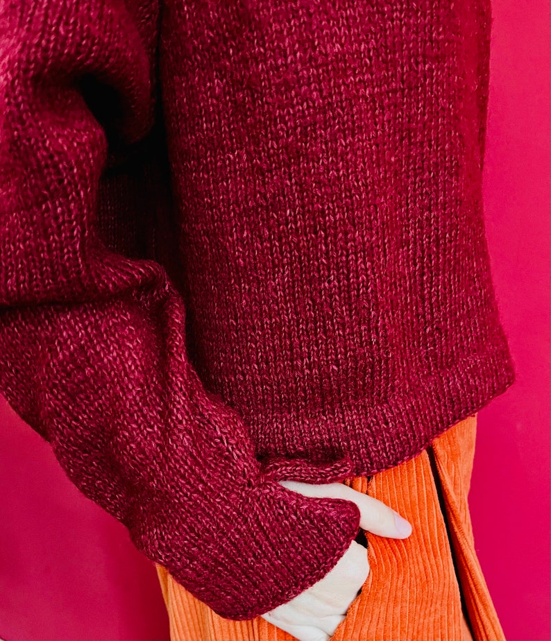 May include: A maroon knitted jumper with long sleeves and a ribbed texture. The jumper is tucked into a pair of orange corduroy trousers.
