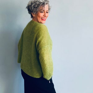 Könnte beinhalten: Eine Frau trägt einen grünen, flauschigen Pullover und eine schwarze Hose. Sie lächelt und schaut über ihre Schulter.