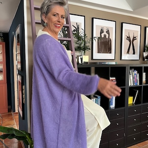 Könnte beinhalten: Eine Frau trägt einen langen, lila Cardigan über einem weißen Oberteil. Sie steht auf einer Leiter vor einem Bücherregal mit Kunstdrucken an der Wand hinter ihr.