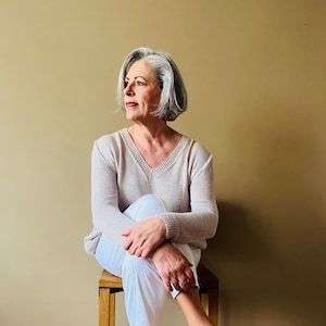 Könnte beinhalten: Eine Frau mit grauem Haar sitzt auf einem Holzhocker. Sie trägt einen hellrosa Pullover und eine weiße Hose. Sie schaut nach rechts.