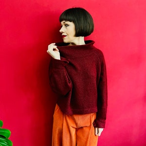 May include: A woman wearing a maroon turtleneck sweater with a ribbed texture and orange corduroy trousers. She is standing in front of a red background.