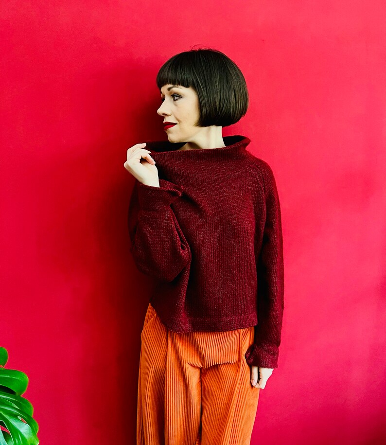 May include: A woman wearing a maroon turtleneck sweater with a ribbed texture and orange corduroy trousers. She is standing in front of a red background.