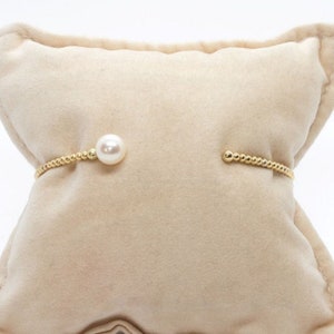 May include: A gold-toned bracelet with a single white pearl and a beaded design, displayed on a beige velvet pillow. The bracelet has an open design. The background is white.
