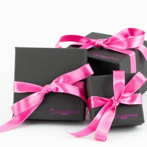 Puede incluir: Tres cajas de regalo negras con lazos de cinta de sat&eacute;n rosa. Las cajas est&aacute;n apiladas una encima de la otra. La caja superior tiene el texto "CusinaBijoux" impreso.