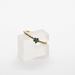 Puede incluir: Un anillo de oro con un dise&ntilde;o de flor verde y blanca. El anillo est&aacute; en un expositor transparente.