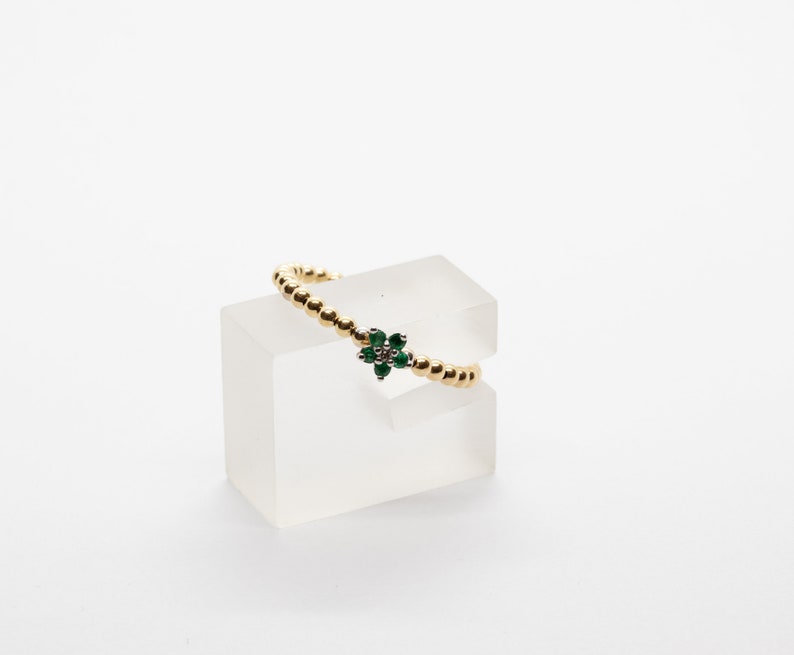 Puede incluir: Un anillo de oro con un dise&ntilde;o de flor verde y blanca. El anillo est&aacute; en un expositor transparente.