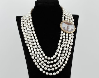 May include: A multi-strand pearl necklace with a cameo brooch featuring a bridge design. The necklace is made of white pearls and the brooch is gold-toned with a brown cameo.