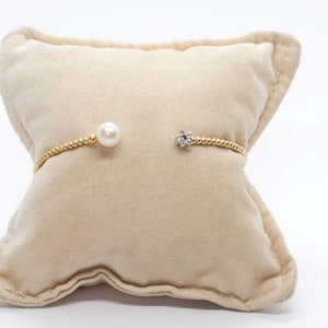 Puede incluir: Un brazalete de oro con una perla y un pequeño dije en forma de flor con piedras transparentes. El brazalete se muestra sobre un cojín de terciopelo beige.