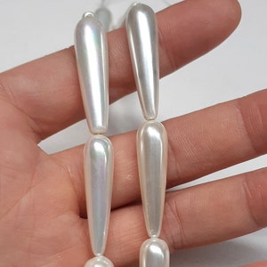 May include: A strand of white pearlescent teardrop-shaped beads. The beads are strung on a clear thread and have a smooth, glossy finish.