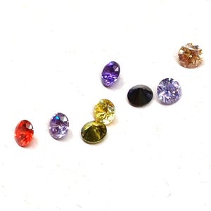 Pode incluir: Uma coleção de pedras preciosas coloridas em várias formas e tamanhos. As pedras incluem vermelho, roxo, amarelo, verde, preto e castanho. As pedras preciosas estão espalhadas sobre uma superfície branca.