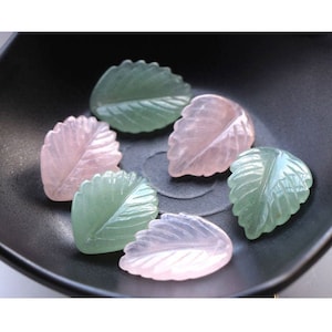 May include: Six carved leaf-shaped pendants made of pink and green gemstone. The leaves are arranged in a circle on a black surface.