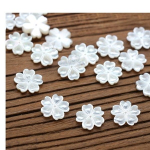 Puede incluir: Una colección de pequeños adornos blancos con forma de flor. Cada flor tiene cinco pétalos y un brillo nacarado. Los adornos están dispersos sobre una superficie de madera, creando un efecto delicado y decorativo.