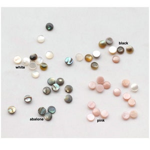 Puede incluir: Surtido de cabujones redondos de concha en varios colores, incluyendo blanco, negro, rosa y abulón. Los cabujones están dispuestos en grupos por color sobre una superficie blanca.