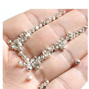 May include: A close-up of a hand holding a pile of silver-colored, faceted beads. The beads are small and have a geometric shape.