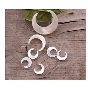 May include: A set of six white mother of pearl crescent moon shaped charms. The charms are of varying sizes and are arranged on a wooden surface.