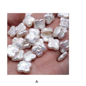 May include: A collection of white flower-shaped pearl beads. The pearls have an iridescent sheen and are arranged in a pile on a hand.