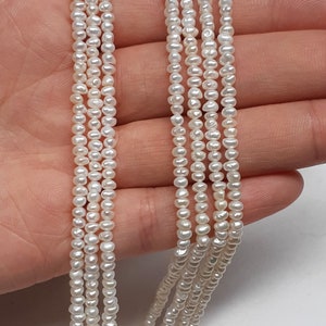 Op de afbeelding: Een streng witte zoetwaterparels, ongeveer 3 mm groot, is te zien op de afbeelding. De parels zijn geregen op een dun draad en zijn in een rechte lijn gerangschikt. De parels zijn glad en hebben een licht iriserende glans.