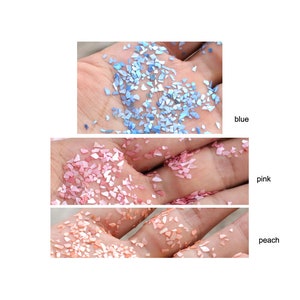 May include: Three different colours of iridescent glitter flakes in a hand. The colours are blue, pink, and peach.