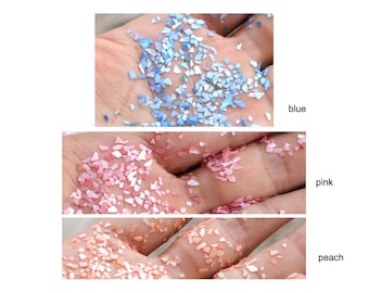 May include: Three different colors of iridescent glitter flakes in a hand. The colors are blue, pink, and peach.