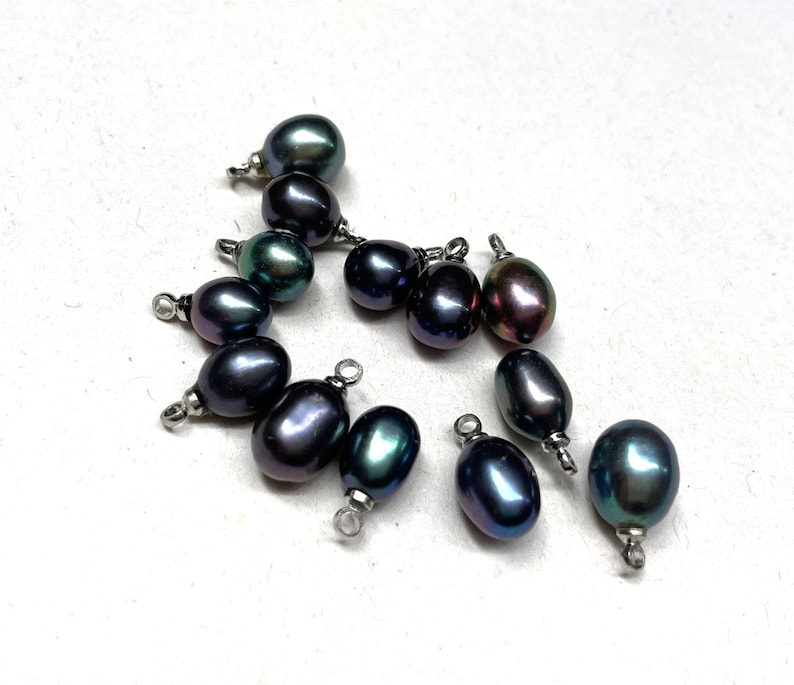May include: A group of 11 dark green and black pearl beads with silver jump rings. The pearls are oval shaped and have a slight iridescent sheen.