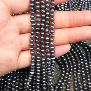 May include: A strand of black freshwater pearls, with a slight iridescent sheen, is displayed on a hand. The pearls are round and uniform in size.