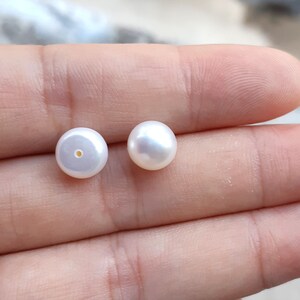 May include: Two white round pearls, one with a hole drilled through the center, on a hand.