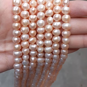 May include: Strands of lustrous, peach-colored pearls. The pearls are round and have a smooth, slightly irregular surface. The strands are held in a hand, showcasing the color and texture of the pearls. Ideal for jewelry making.