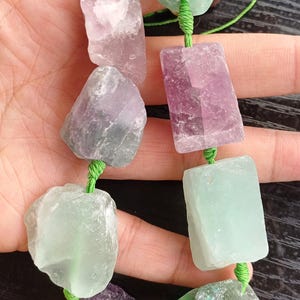 Puede incluir: Un collar con piedras de fluorita en bruto multicolores. Las piedras, en tonos morados, verdes y claros, están ensartadas en un cordón verde. Las piedras, de formas y tamaños variados, tienen un aspecto natural y sin pulir. El collar se sostiene en una mano.