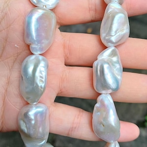 Pode incluir: Um fio de pérolas brancas grandes, irregulares e brilhantes. As pérolas têm um brilho iridescente, com tons de rosa e azul. As pérolas são enfiadas, possivelmente formando um colar ou pulseira.