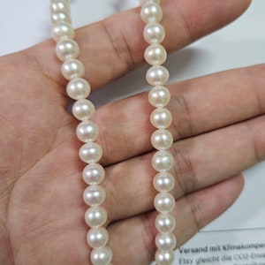 Pode incluir: Um fio de pérolas brancas, enfiadas em um fio fino, é segurado em uma mão. As pérolas são redondas e lisas, e o fio tem cerca de 46 cm de comprimento.