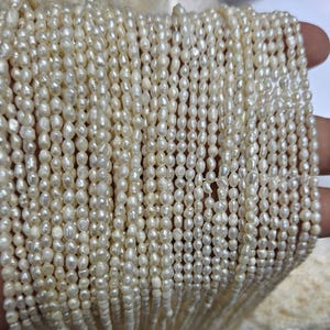 May include: Close-up of strands of creamy white, oval-shaped pearls. The pearls are strung together, creating a dense, textured surface. The pearls vary slightly in size and shape, reflecting natural variations.