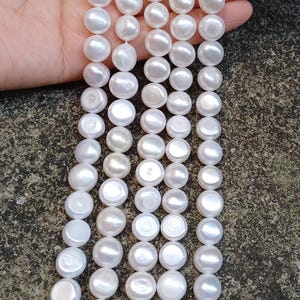 Puede incluir: Una hebra de perlas de agua dulce blancas, cada perla es redonda y lisa. Las perlas están enhebradas en un hilo blanco y están dispuestas en línea recta. Las perlas tienen un diámetro aproximado de 8 mm.