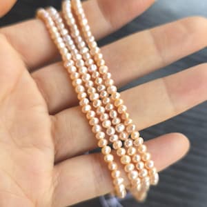 Puede incluir: Cuatro hilos de delicadas perlas color melocotón se sostienen en una mano. Las perlas son pequeñas y redondas, ensartadas con un hilo fino. Los hilos tienen longitudes ligeramente diferentes, creando un efecto en capas. Las perlas tienen un brillo sutil.