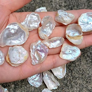 May include: A collection of white, iridescent, natural, irregular shaped, pearl shell, with a hole drilled through each one. The shells are on a hand, and appear to be for jewellery making.