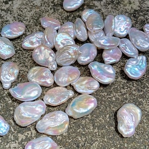 Pode incluir: Uma coleção de pérolas iridescentes de formato irregular. As pérolas têm uma cor de base branca perolada e brilhante com tons brilhantes de rosa, azul e verde. As pérolas estão sobre uma superfície texturizada cinza escuro.