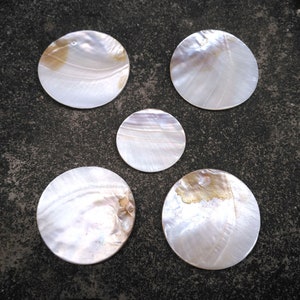Może przedstawiać: Pięć okrągłych, białych podkładek z perłowej muszli z opalizującymi wzorami. Podkładki są gładkie i wypolerowane.