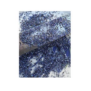 May include: A collection of small, iridescent dark blue crushed stone fragments scattered across a surface. A metal ruler is placed over the stones, showing markings that indicate a measurement range between 15 and 21 units in length.