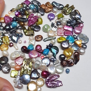 Op de afbeelding: Een verzameling van iriserende en parelmoerachtige kralen in verschillende vormen en maten, waaronder ronde, ovale en onregelmatige vormen. De kralen zijn in tinten wit, grijs, roze, paars, blauw, groen en goud.