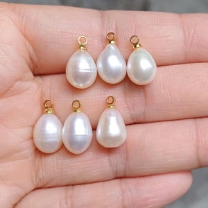 May include: Six white pearl teardrop charms with gold-toned jump rings. The charms are displayed on a hand.