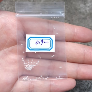 Könnte beinhalten: Ein kleiner transparenter Plastikbeutel gefüllt mit winzigen weißen Perlen. Der Beutel trägt ein Etikett mit der Aufschrift "0,9 mm".