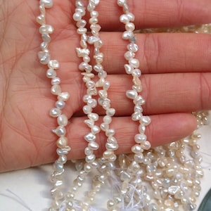 Può includere: Un filo di perle d'acqua dolce bianche, con forme e dimensioni irregolari, infilate su un filo bianco. Le perle sono disposte in un motivo sciolto e fluido.