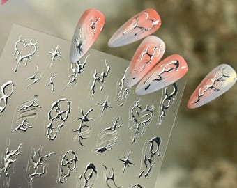 Puede incluir: Una hoja de pegatinas adhesivas para uñas con diseños metálicos plateados de estilo líquido, que incluyen corazones derretidos, estrellas y formas orgánicas abstractas. Varios adhesivos están aplicados sobre uñas postizas.