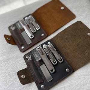 May include: Two open brown leather manicure kits featuring silver metal tools. Each kit contains a nail file, three nail clippers, and a small pick tool. The tools are engraved with the word MANICURE. The leather cases have snap button closures.