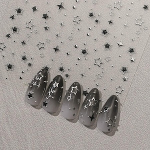 May include: A set of five almond-shaped press-on nails featuring a black-to-white gradient finish. Each nail is decorated with metallic silver star-shaped decals and small dots. A matching sheet of silver star and dot nail stickers is included.