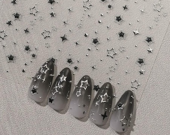 May include: A set of five almond-shaped press-on nails featuring a black-to-white gradient finish. Each nail is decorated with metallic silver star-shaped decals and small dots. A matching sheet of silver star and dot nail stickers is included.